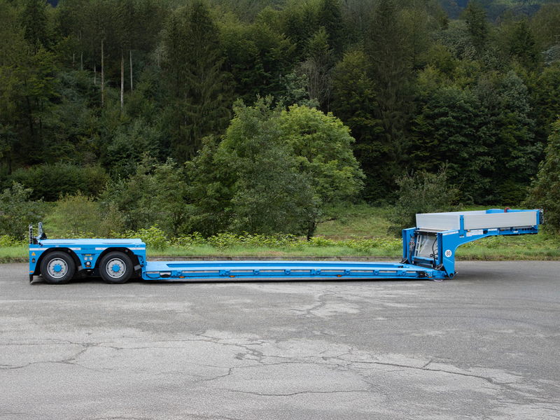 Blue low-loader trailer with hydraulic ramps, designed for transporting heavy equipment, parked on a roadside.