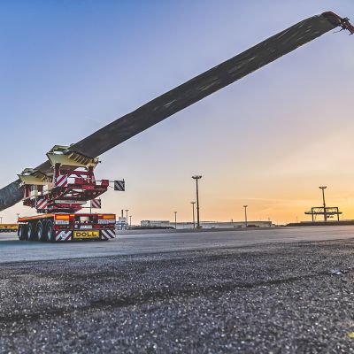 A DOLL heavy-duty trailer transports a wind turbine blade at a port site during sunset.