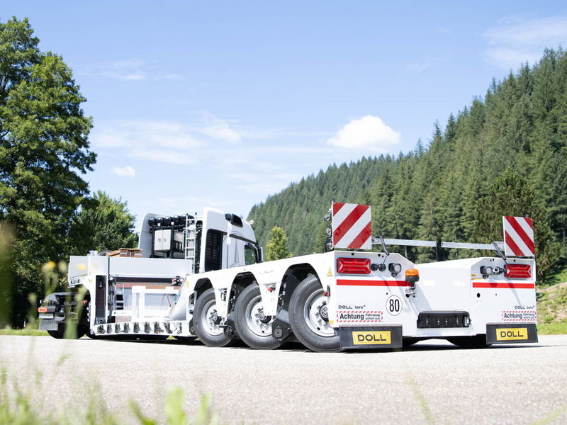 White low-bed trailer attached to a truck, equipped for heavy transport, parked on a road near a forest.