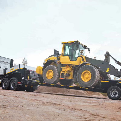Semi-remorque surbaissée DOLL vue de côté, chargée d'un véhicule de chantier, sur un sol sablonneux