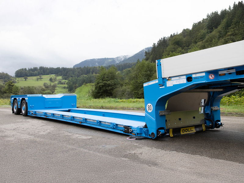 Blue low-loader trailer with hydraulic ramps, designed for transporting heavy equipment, parked on a roadside.