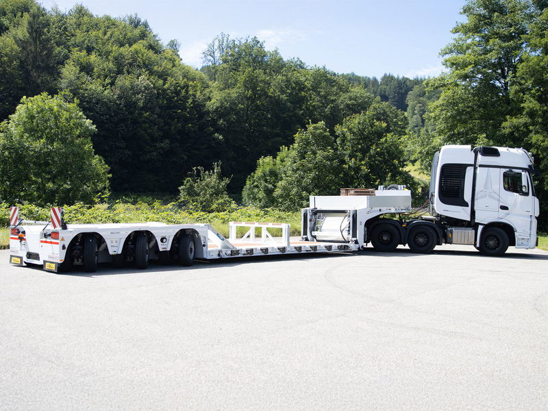 White low-bed trailer attached to a truck, equipped for heavy transport, parked on a road near a forest.
