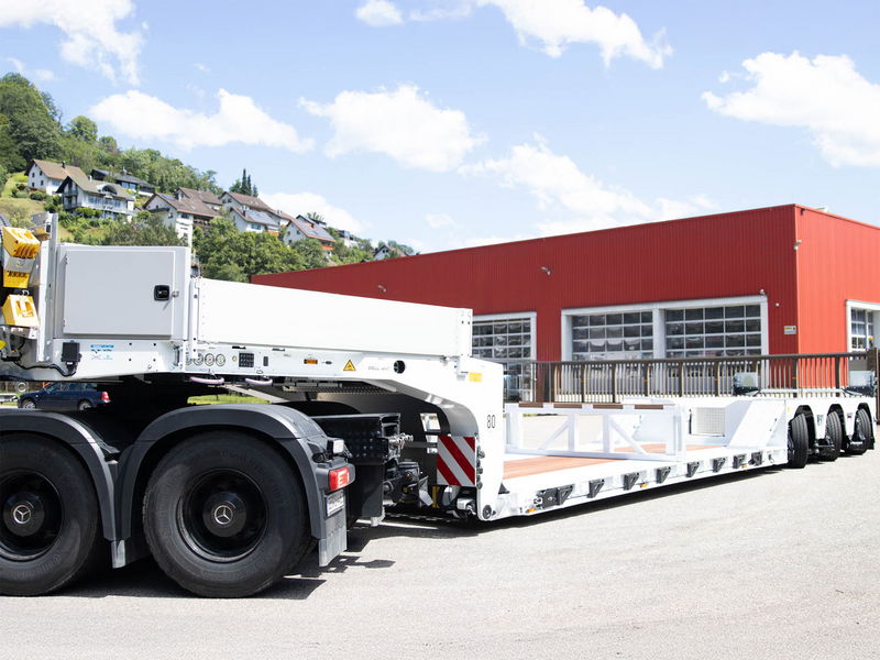 Large white low-bed trailer connected to a Mercedes-Benz truck, parked near a red warehouse.