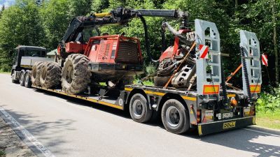 Heavy-duty tractor secured on a flatbed trailer for transportation through a forested road under a sunny sky.